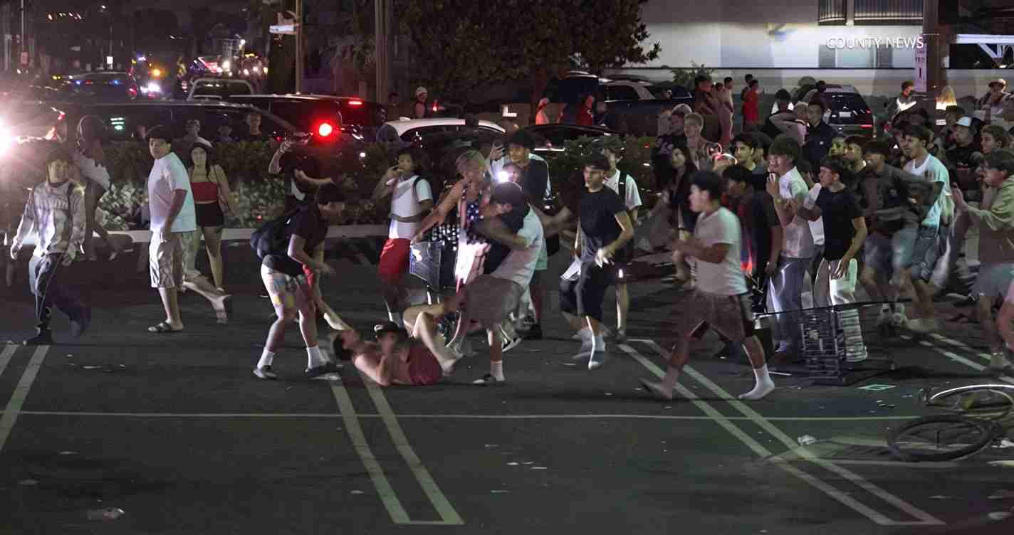 Nighttime urban street scene with a large crowd of pedestrians; some appear to be involved in a scuffle while a person lies on the pavement in the center.