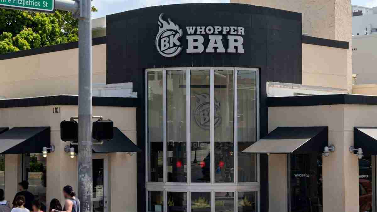 Front view of a BK Whopper Bar storefront with a large curved glass entrance and black signage reading 'WHOPPER BAR' above. Street corner with people nearby.
