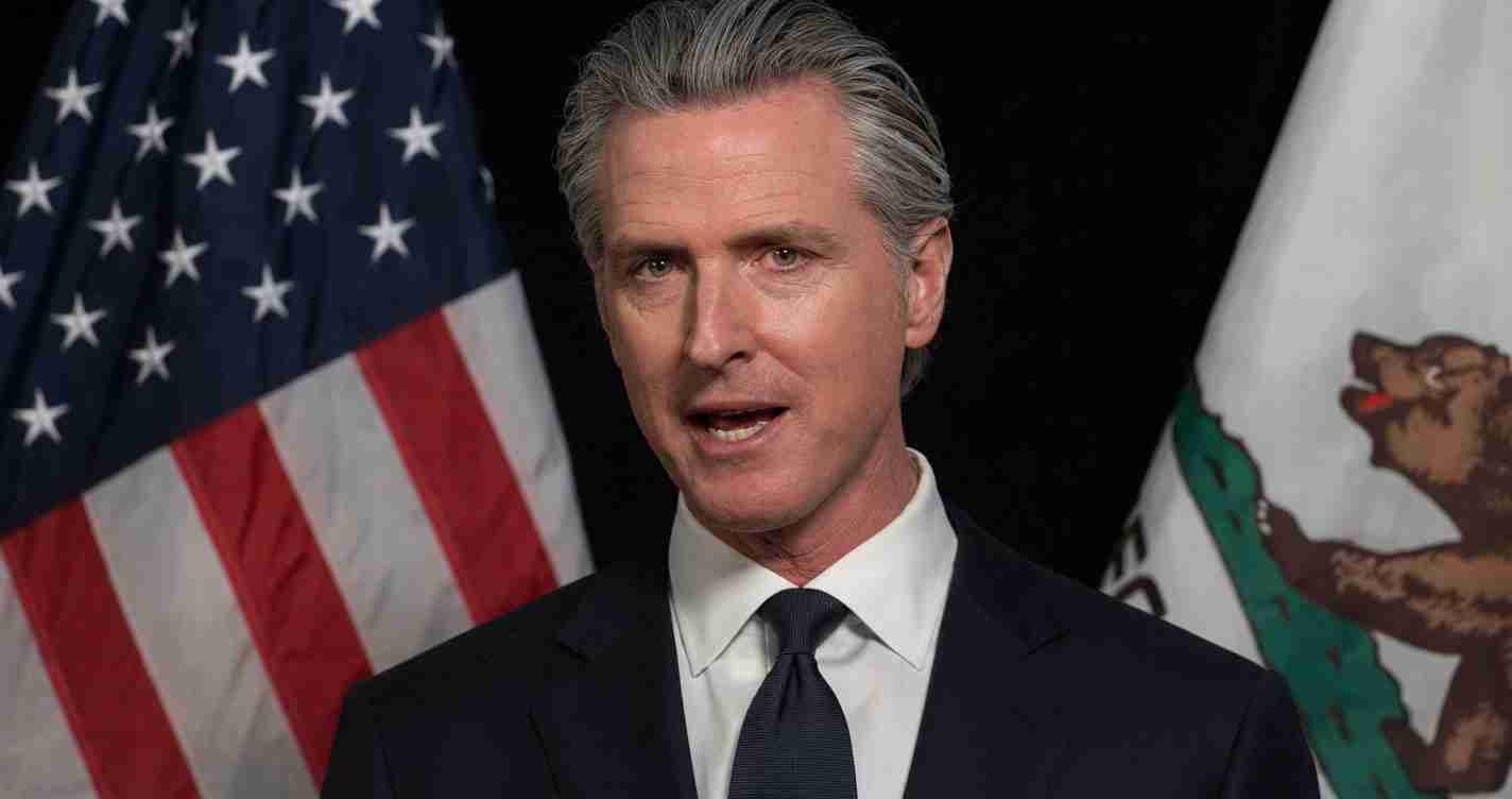 Man in a dark suit speaking, with U.S. flag and another flag in the background behind him.