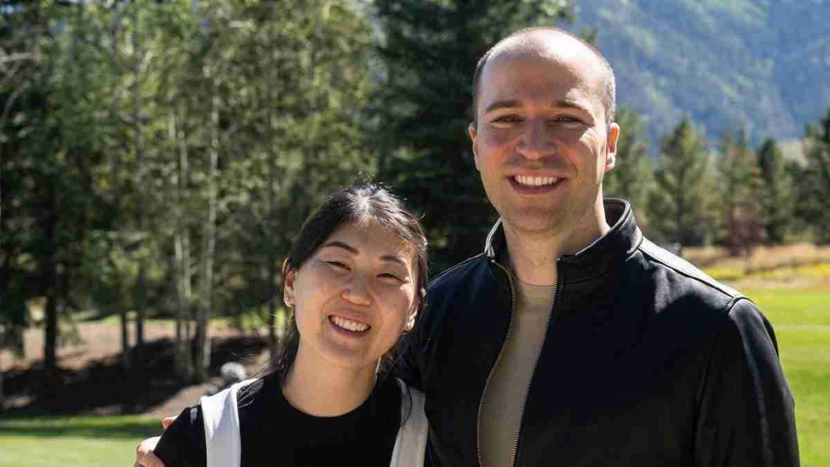 Smiling man and woman standing together outdoors in a park with trees and distant hills in the background.