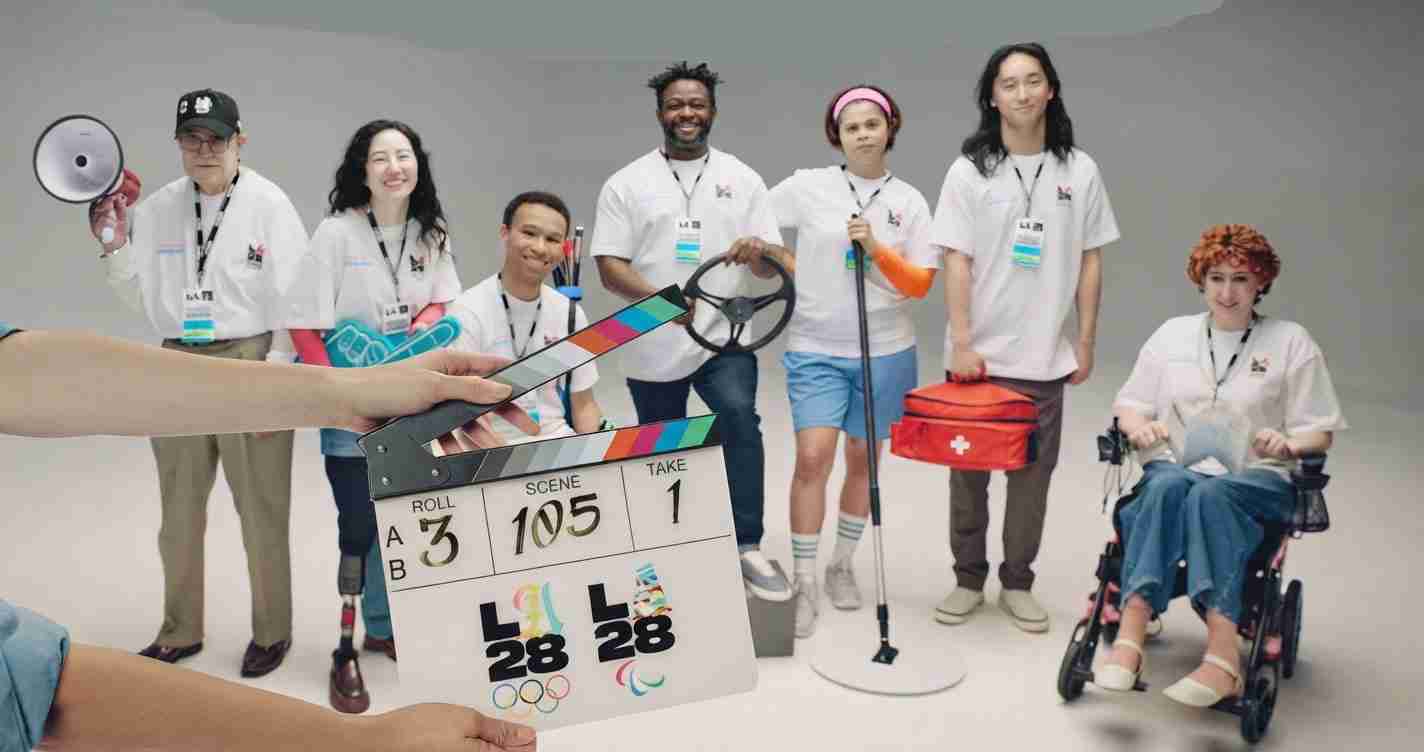 Diverse group of volunteers in white shirts pose in a studio; a clapperboard is held foreground with a person in a wheelchair on the right and a first-aid bag nearby.