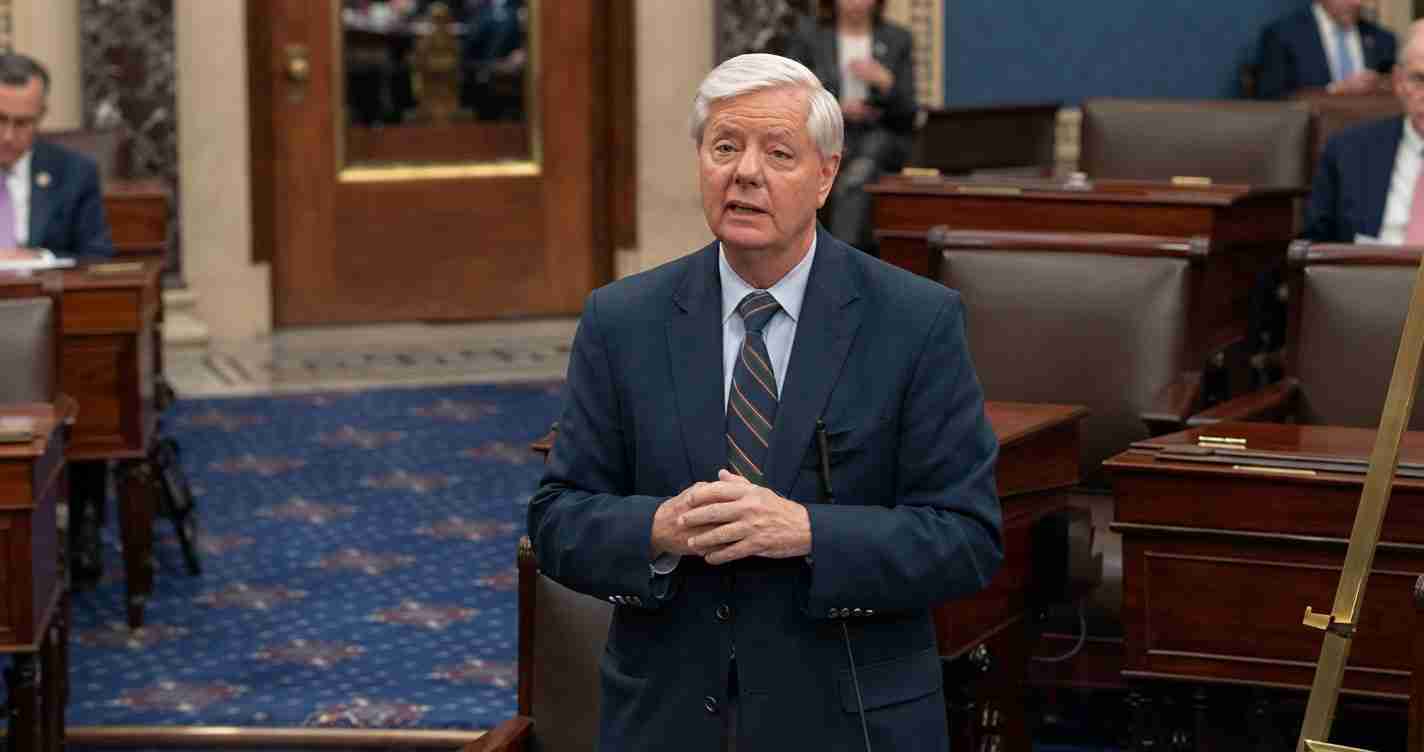 Man with white hair in a navy suit speaks in a legislative chamber, hands clasped in front of him.