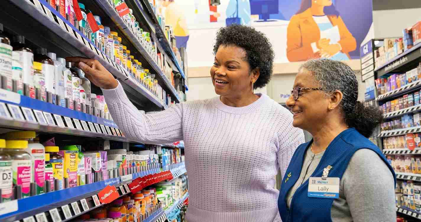 Two women in a store aisle smile as one selects a product from the shelf nearby the others look on happily.