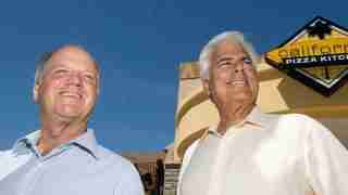 Two smiling men stand outside a California Pizza Kitchen, blue sky behind them.