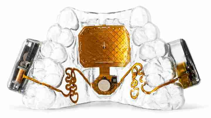 Cross-section model of an implantable medical device with an orange circuit board embedded in a clear dental arch model. The central orange module has a cross pattern and is connected to gold-tone circuitry along the sides.